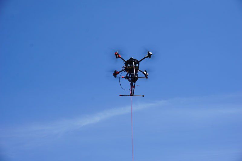 Multikopter-Drohne mit Seilverbindung schwebt vor blauem Himmel während eines Einsatzes oder Trainings.