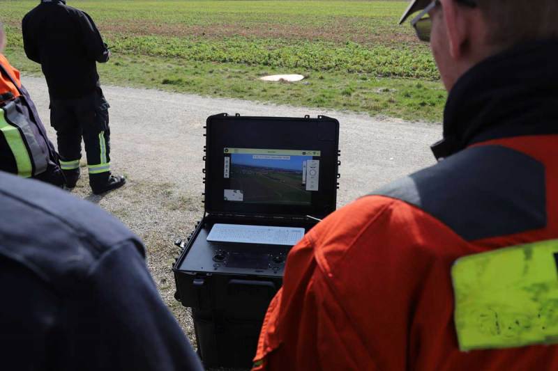 Personen in Einsatzkleidung betrachten die Live-Übertragung einer Drohnenkamera auf einem mobilen Monitor im Feld.